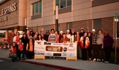 A large group of Many Parts, One Body volunteers, students, and supporters gathered outside Blue Springs High School at sunset, holding the organization's banner ringed with sponsor logos
