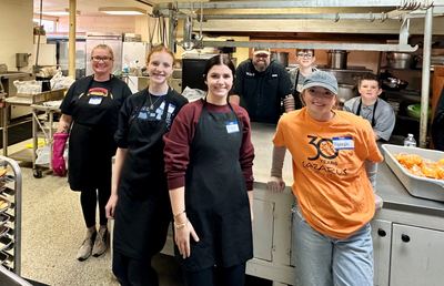 Many Parts, One Body volunteers in aprons preparing trays of hot food in a community kitchen